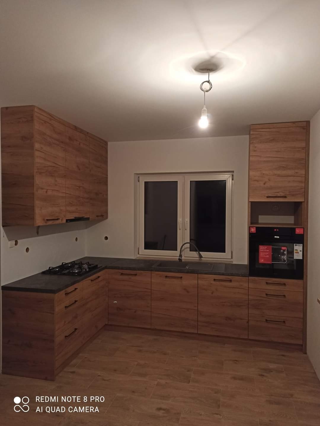 Nowoczesna zabudowa kuchenna z drewnianymi frontami i czarnym blatem w domu w Prokowie. Widoczna płyta grzewcza, zlew i piekarnik. Uwagę zwraca wisząca żarówka bez klosza.