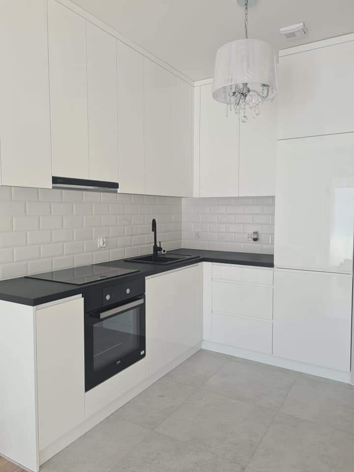 Jasna kuchnia w stylu minimalistycznym z białymi frontami szafek, czarnym blatem i armaturą oraz białymi płytkami ściennymi imitującymi cegiełki, oświetlona ozdobnym żyrandolem.