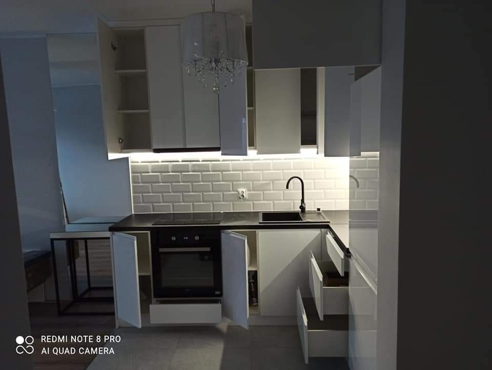 Nowoczesna, biała kuchnia z czarnym blatem i armaturą, oświetlenie LED pod szafkami, otwarte szafki i szuflady, widoczna biała cegła na ścianie.