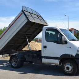 Biały samochód dostawczy z otwartą skrzynią załadowaną piaskiem, podniesioną hydraulicznie do góry, widoczny na parkingu w słoneczny dzień.
