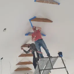 Dwóch mężczyzn na rusztowaniu maluje biały sufit z drewnianymi elementami, zabezpieczonymi folią i taśmą malarską, widoczne kable i przygotowane otwory na oświetlenie.