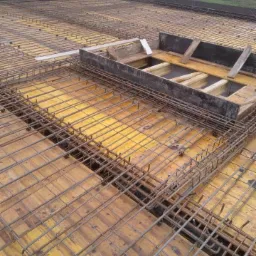 Zbrojenie stropu z deskowaniem i otworem na schody, widok z góry na plac budowy w pochmurny dzień.