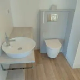 Nowoczesna łazienka z okrągłą umywalką nablatową na blacie z szarego kamienia i podwieszanym WC z zabudową w odcieniach szarości, podłoga wyłożona panelami imitującymi drewno.