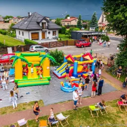 Rozległy plac zabaw z dmuchanymi zamkami, zjeżdżalniami i basenem z piłeczkami, otoczony bawiącymi się dziećmi i dorosłymi, widziany z góry, z budynkami w tle, w tym sklep z lodami 'Consonni Lody...