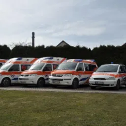 Flota pojazdów ratunkowych, w tym ambulanse i samochody osobowe z oznaczeniami medycznymi, zaparkowana przed zielonym żywopłotem i budynkiem.