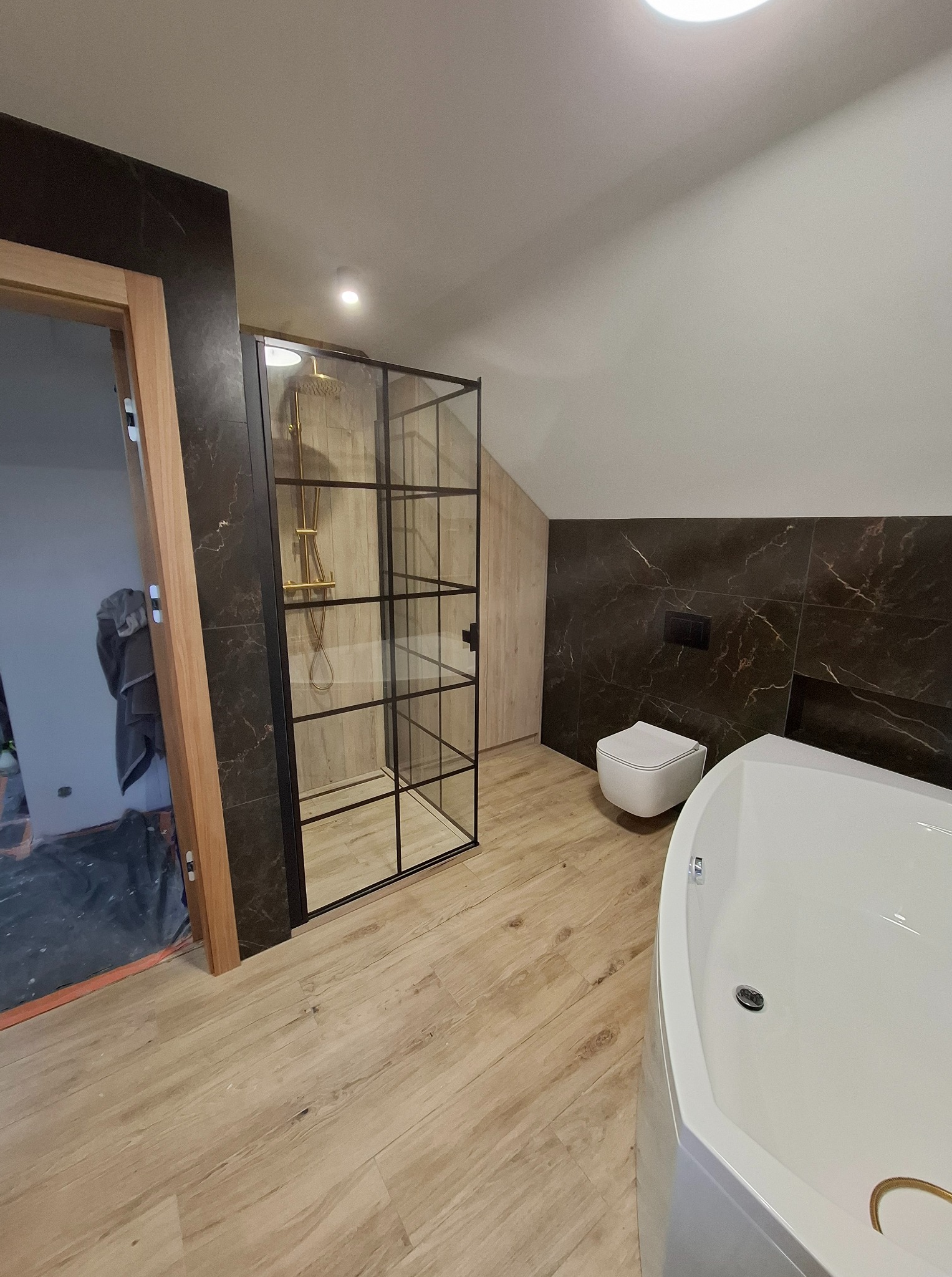 Wykończona łazienka na poddaszu z kabiną prysznicową typu loft, wiszącą toaletą i narożną wanną. Ciemne płytki imitujące marmur na ścianach, jasna podłoga.