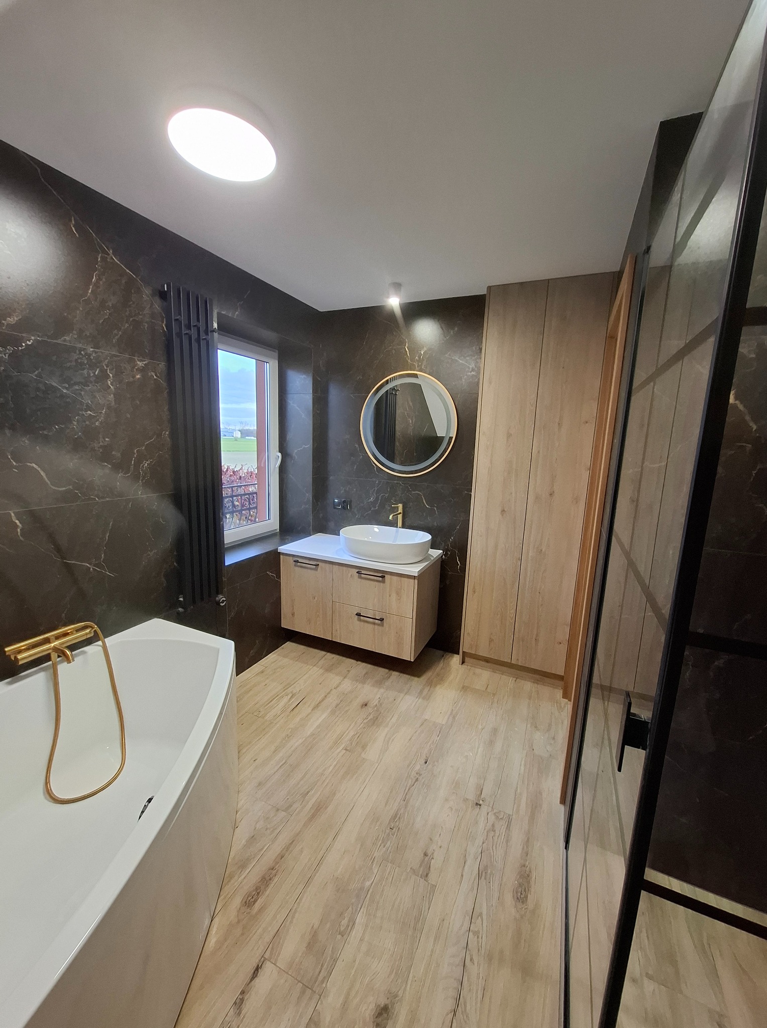 Elegancka łazienka z wanną, umywalką na szafce i kabiną prysznicową. Ciemne marmurowe płytki na ścianach kontrastują z jasną drewnianą podłogą i szafką.