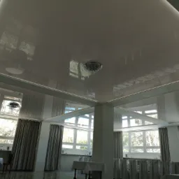 Biały, błyszczący sufit napinany z zamontowanym ozdobnym żyrandolem, odbijający światło z okien w przestronnym pomieszczeniu.