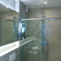 Szklana kabina prysznicowa z deszczownicą i baterią termostatyczną, widoczna umywalka oraz lustro z podświetleniem w nowoczesnej łazience wykończonej beżowymi płytkami.