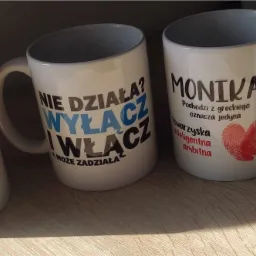 Rząd białych kubków z nadrukami: 'Nie działa? Wyłącz i włącz', 'Monika', imię z koroną i 'Christian / Kebab'.