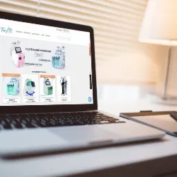 Laptop wyświetlający stronę internetową z ofertą urządzeń do oczyszczania wodorowego, obok tablet graficzny z piórem, w tle lampka stołowa i żaluzje.