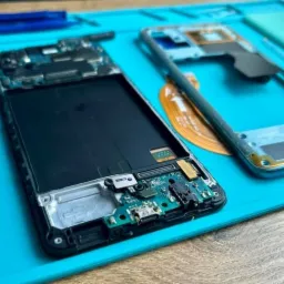 Rozłożony smartfon na niebieskiej macie serwisowej, widoczne elementy elektroniki, obudowa i narzędzia, prawdopodobnie podczas naprawy lub demontażu.