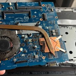 Usługi informatyczne Krystian Widźgowski - Płyta główna laptopa Dell z widocznym układem chłodzenia, wentylatorem i elementami elektronicznymi, trzymana w dłoni nad obudową laptopa. Widoczne detale płyty, takie jak złącza DIMM i oznaczenia...