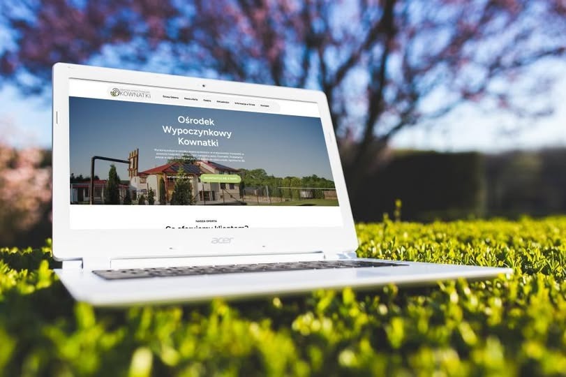 Laptop Acer wyświetlający stronę internetową ośrodka wypoczynkowego Kownatki, umieszczony na trawie z kwitnącymi drzewami w tle, w słoneczny dzień.