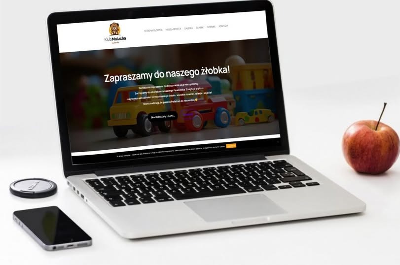 Laptop wyświetla stronę internetową przedszkola z zabawkami w tle, obok leży jabłko, telefon i dekielek obiektywu.