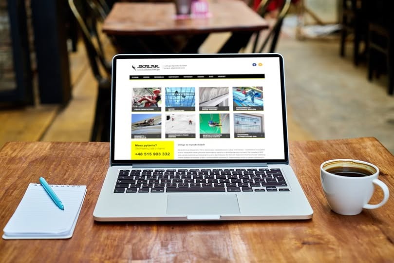 Laptop wyświetla stronę internetową firmy 'Skylar' z galerią zdjęć realizacji montażu i serwisu okien dachowych, leży na drewnianym stole obok notatnika z długopisem i filiżanki kawy.