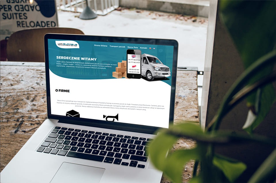 Laptop wyświetlający stronę internetową firmy transportowej Ja-Eurobus z widocznym samochodem dostawczym i numerami telefonów, umieszczony na drewnianym stole z roślinami w tle.