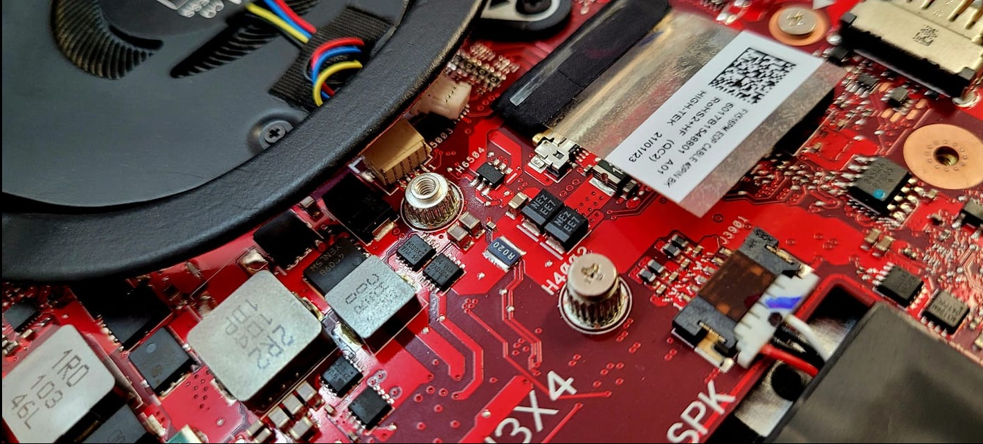 Detale płyty głównej laptopa z widocznym wentylatorem, kondensatorami, układami scalonymi i naklejką z kodem QR. Czerwony laminat płyty głównej z bliska.