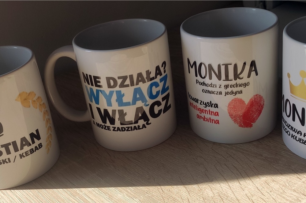 Rząd białych kubków z nadrukami: 'Nie działa? Wyłącz i włącz', 'Monika', imię z koroną i 'Christian / Kebab'.