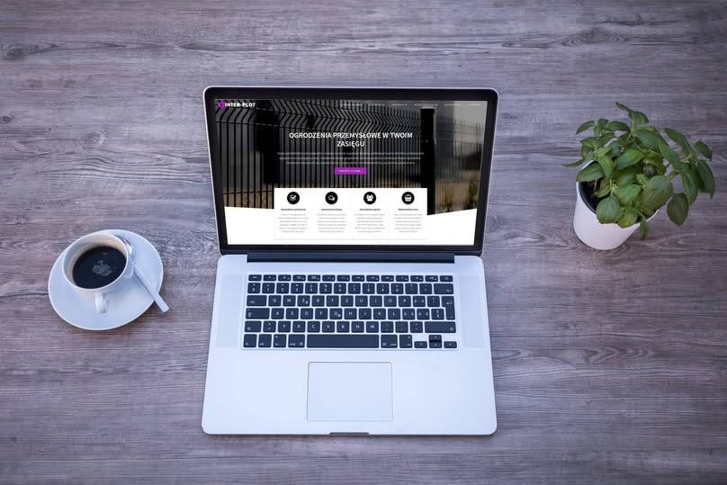 Laptop wyświetlający stronę internetową firmy Inter Plot, zajmującej się ogrodzeniami przemysłowymi, obok filiżanka kawy i mała roślina doniczkowa na drewnianym biurku.
