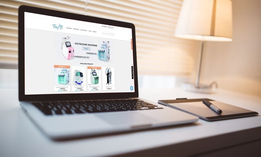 Laptop wyświetlający stronę internetową z ofertą urządzeń do oczyszczania wodorowego, obok tablet graficzny z piórem, w tle lampka stołowa i żaluzje.