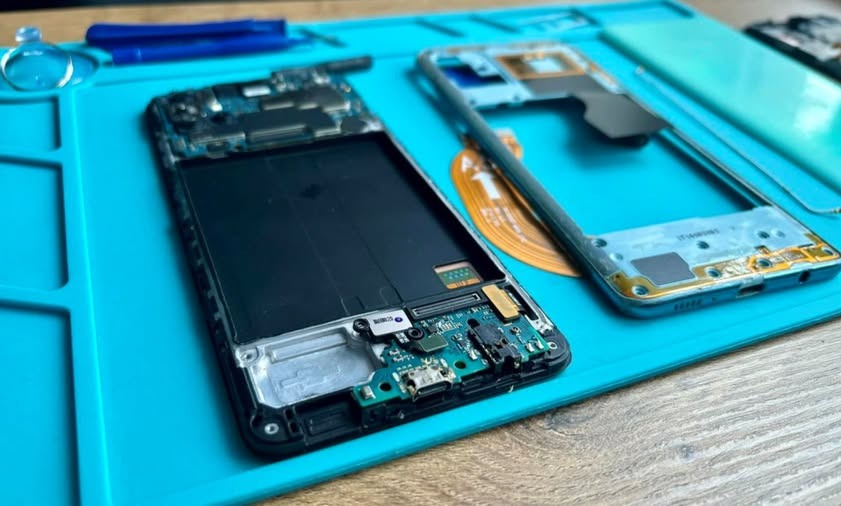 Rozłożony smartfon na niebieskiej macie serwisowej, widoczne elementy elektroniki, obudowa i narzędzia, prawdopodobnie podczas naprawy lub demontażu.