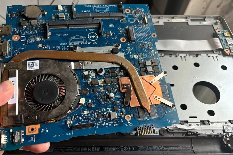 Płyta główna laptopa Dell z widocznym układem chłodzenia, wentylatorem i elementami elektronicznymi, trzymana w dłoni nad obudową laptopa. Widoczne detale płyty, takie jak złącza DIMM i oznaczenia...