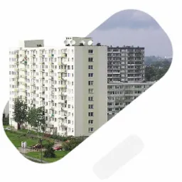 Dwa bloki mieszkalne z balkonami, widoczne anteny satelitarne na dachu wyższego budynku, pochmurne niebo w tle.