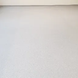 Jasnoszara, gładka podłoga z drobnym wzorem, listwy przypodłogowe w kolorze szarym, grzejniki na ścianie po lewej stronie, jasne ściany, widoczne łączenia paneli podłogowych.