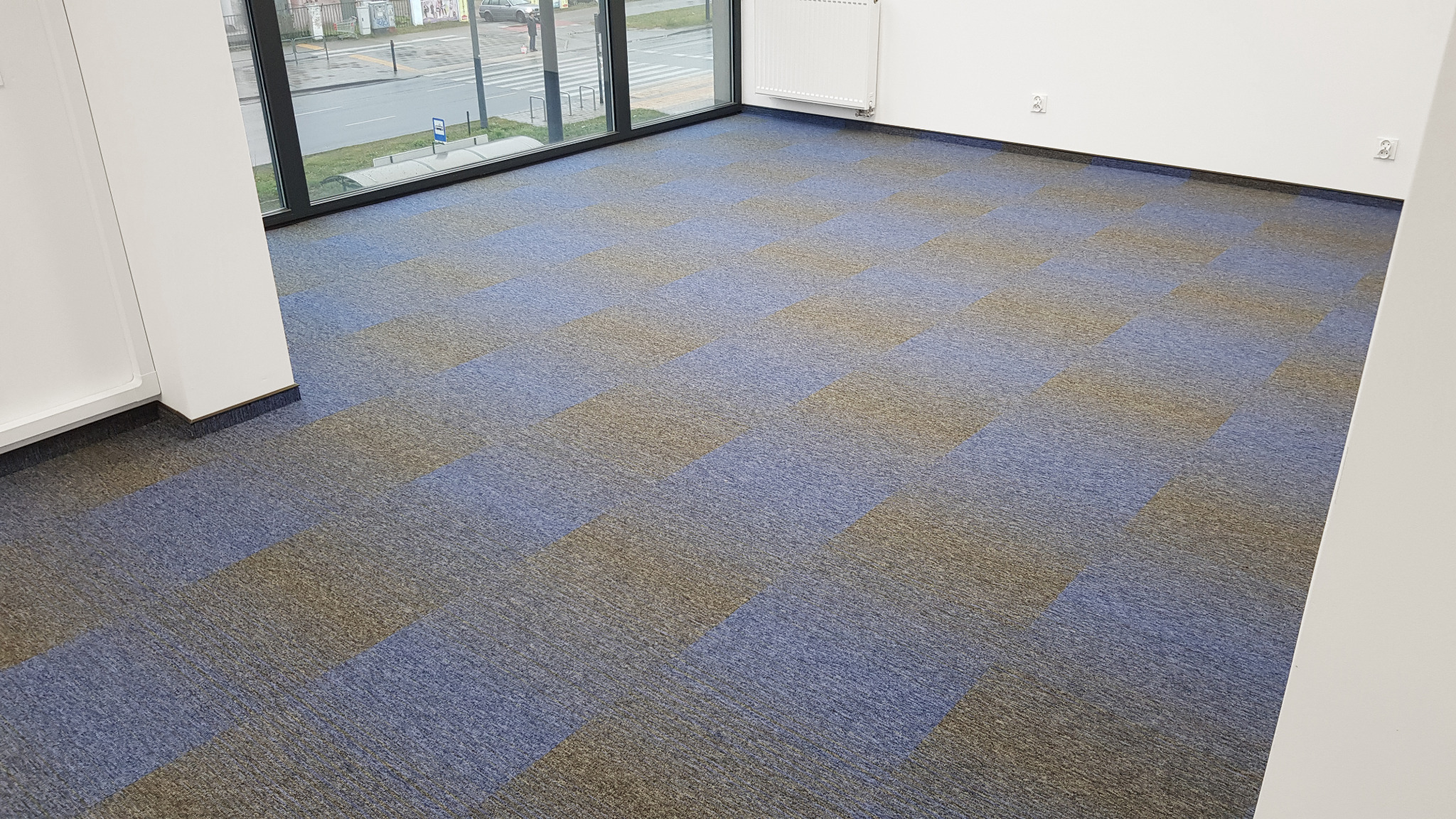 Wykładzina dywanowa w biurze ułożona w szachownicę z kwadratów w odcieniach niebieskiego i szaro-brązowego, widok na okno z ulicą.