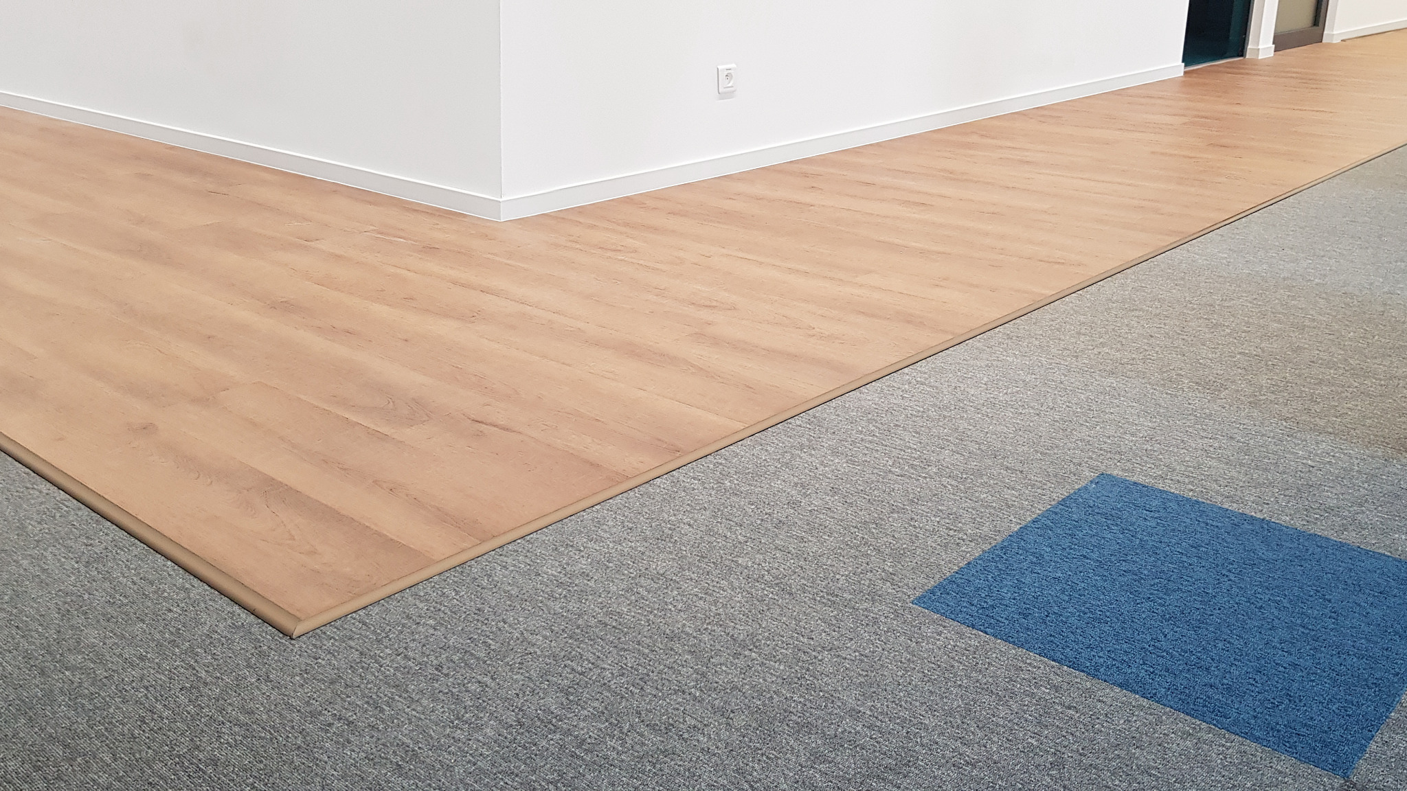 Połączenie podłogi z paneli drewnopodobnych z szarym dywanem w biurze, widoczna listwa przypodłogowa i fragment ściany z gniazdkiem.