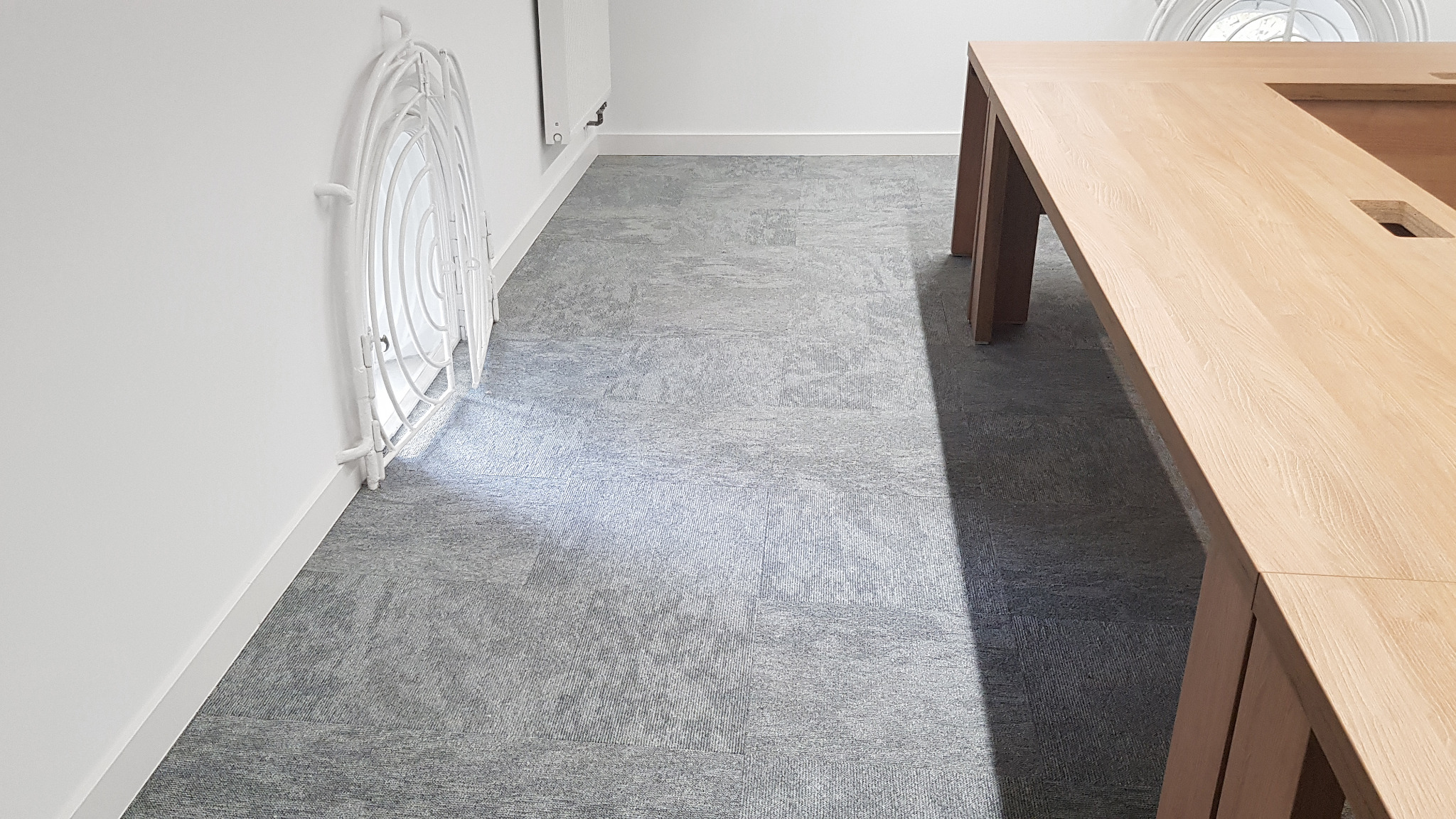 Jasnoszara wykładzina dywanowa w biurze, widoczny fragment drewnianego stołu konferencyjnego i biały grzejnik za dekoracyjną osłoną.