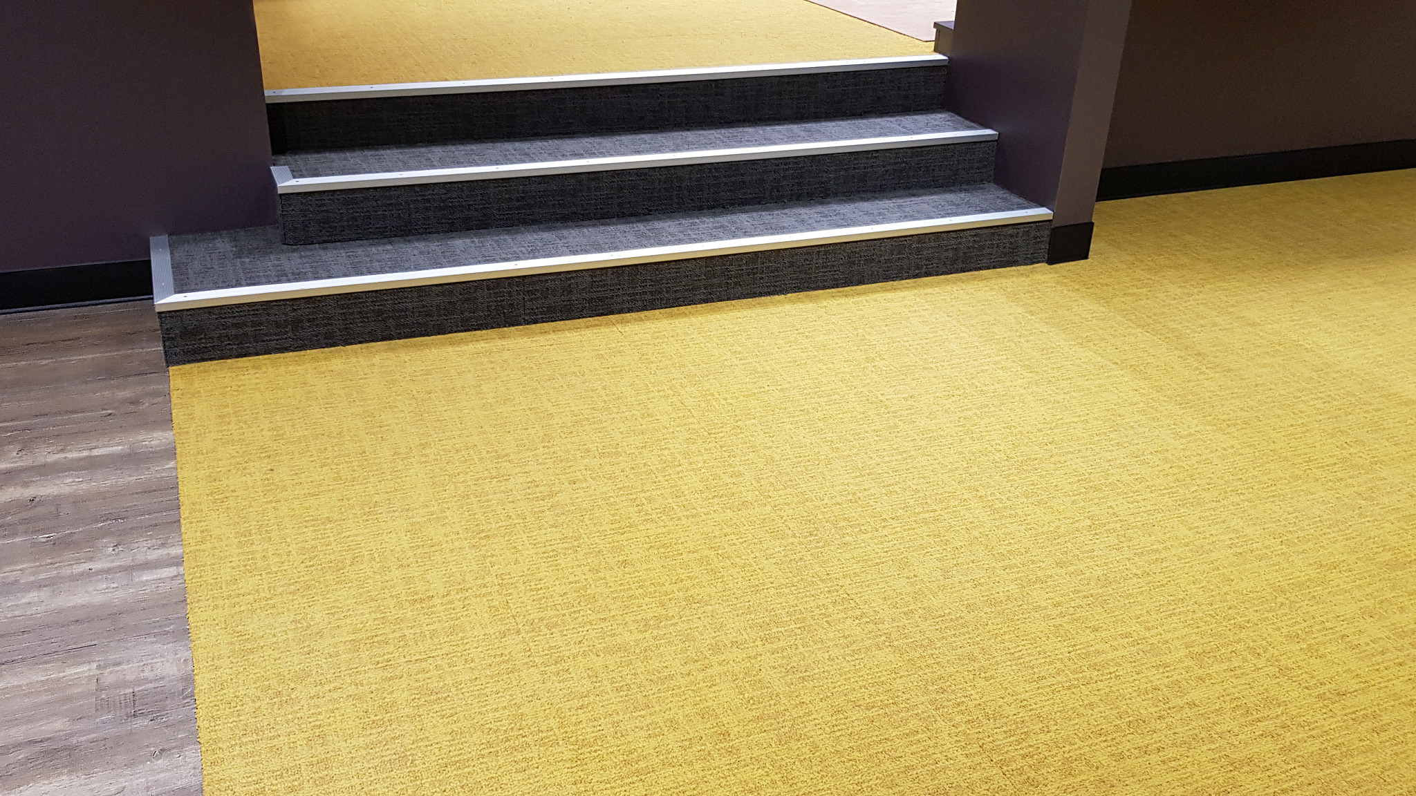 Żółta wykładzina dywanowa w biurze, schody z szarą wykładziną i aluminiowymi listwami krawędziowymi, ściany w odcieniach fioletu i brązu.