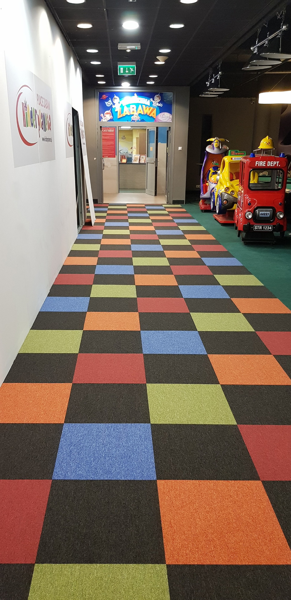 Kolorowa wykładzina podłogowa w szachownicę w korytarzu placu zabaw, widoczne zabawki-pojazdy i wejście do strefy zabawy.