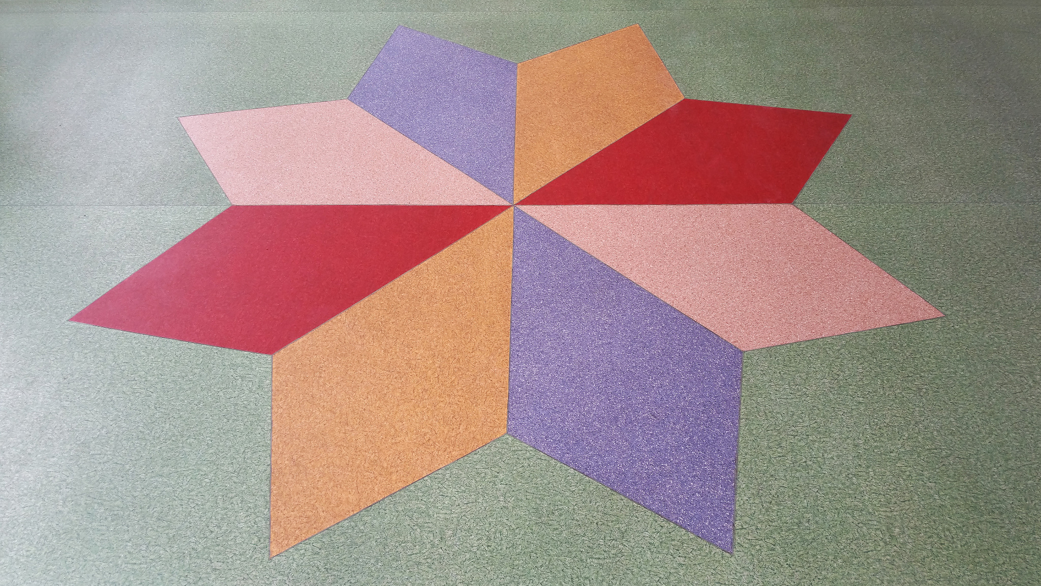 Wielobarwna, ośmioramienna gwiazda ułożona z trójkątnych elementów na podłodze o zielonym, teksturowanym tle. Elementy gwiazdy w kolorach: czerwonym, pomarańczowym, różowym i fioletowym.