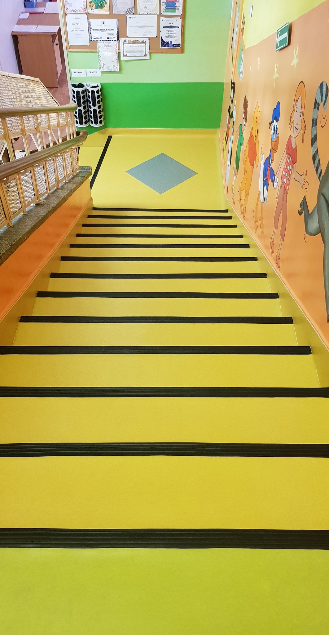 Schody w przedszkolu z żółtymi stopniami i czarnymi paskami antypoślizgowymi, ściany pomalowane w jasne kolory z motywami bajkowymi.