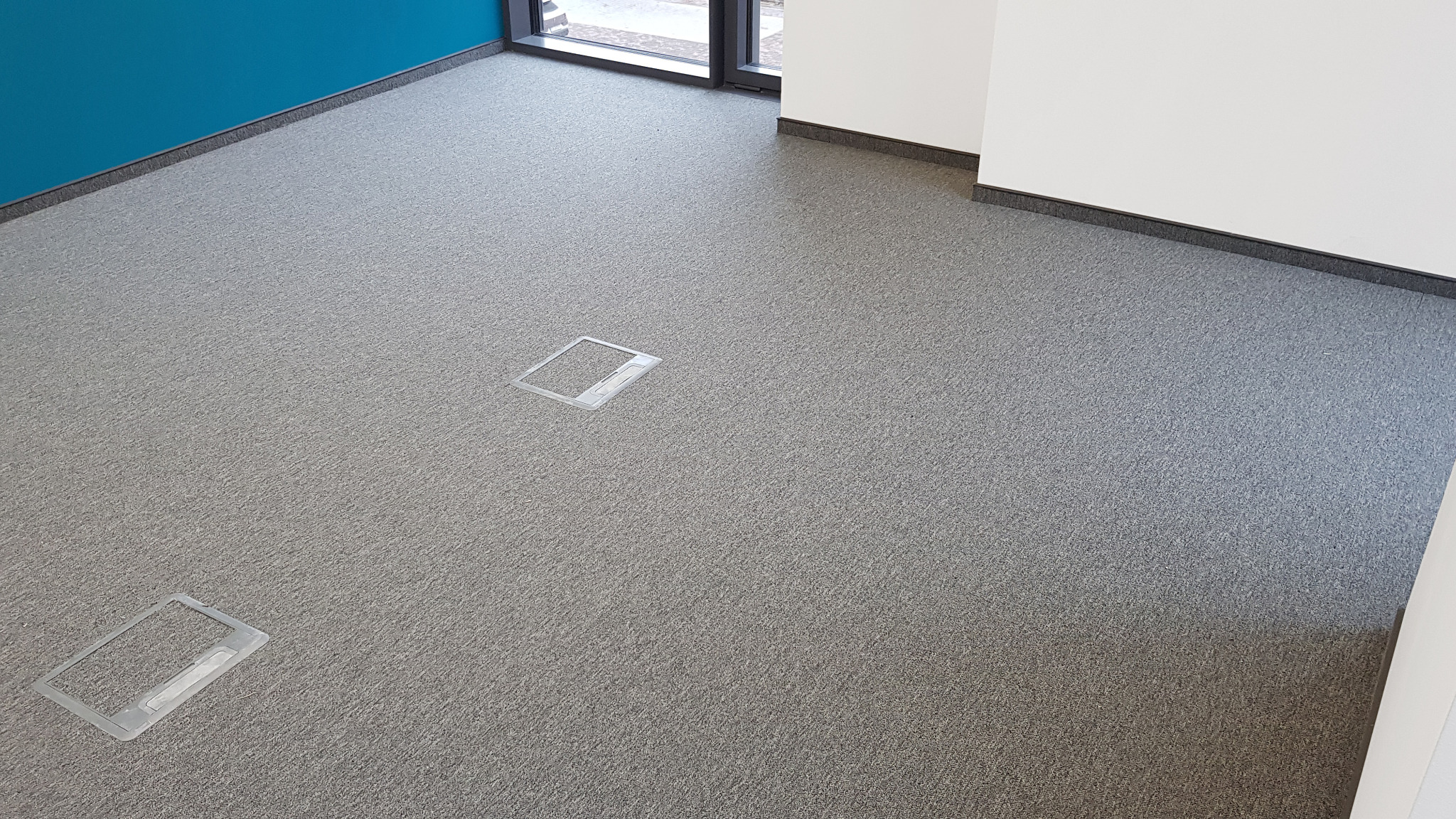 Wnętrze biura z szarą wykładziną dywanową i dwoma gniazdami podłogowymi z przezroczystymi klapkami, ściana pomalowana na niebiesko w rogu pomieszczenia.