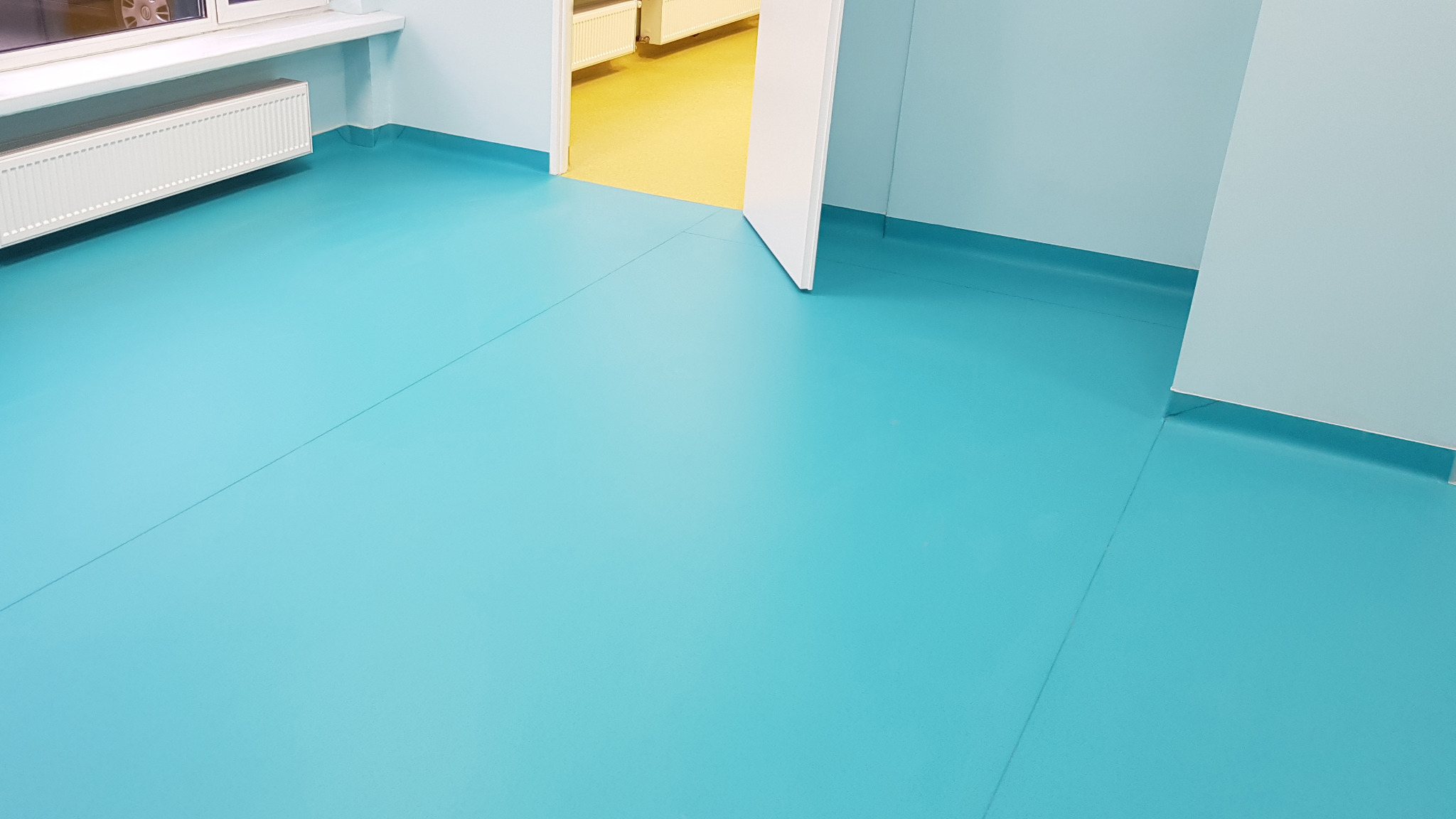 Turkusowa podłoga z PVC w pomieszczeniu z jasnoniebieskimi ścianami i białym grzejnikiem, widoczny fragment żółtej podłogi w sąsiednim pomieszczeniu przez otwarte drzwi.