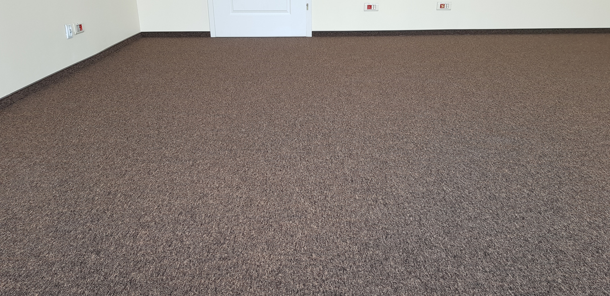 Nowa, brązowa wykładzina dywanowa w pokoju, widoczne gniazdka elektryczne i białe drzwi w tle.