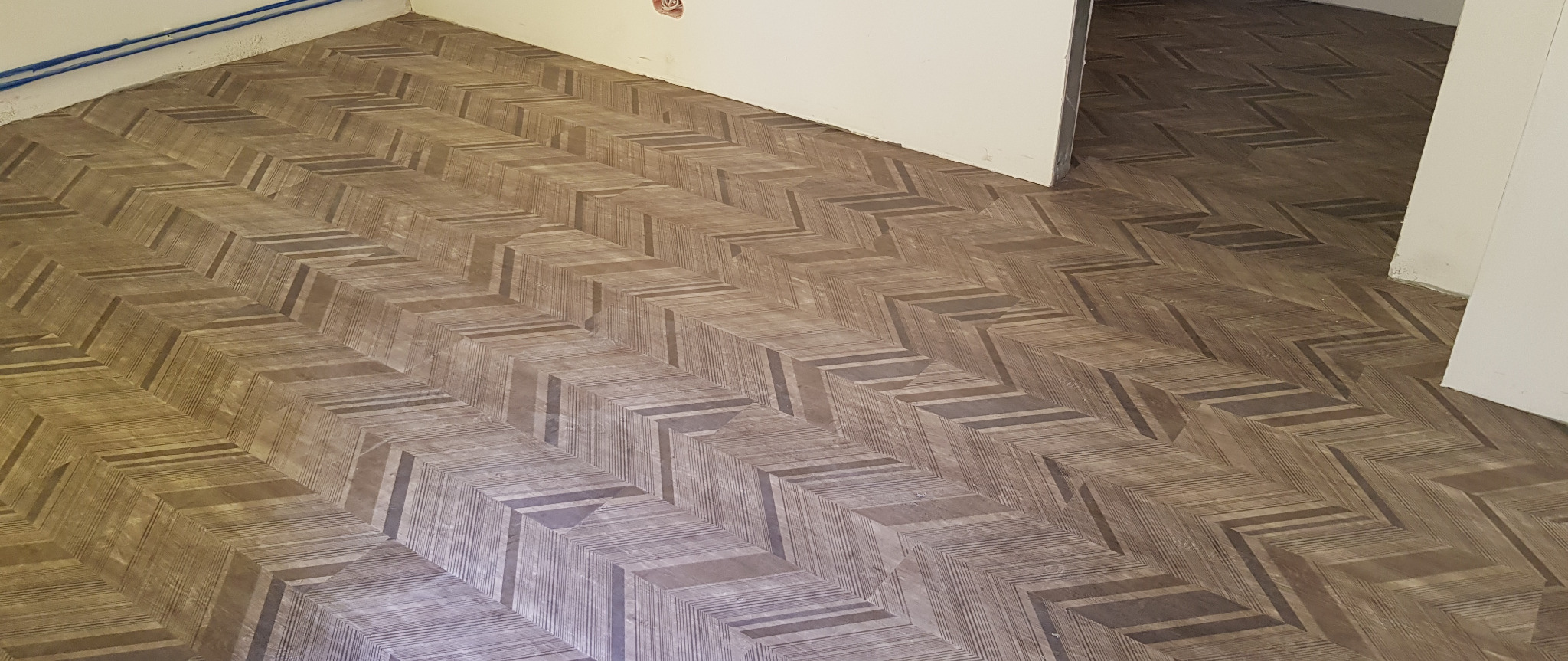 Ułożona w jodełkę, brązowa podłoga z paneli z widoczną strukturą drewna, w nowym, jasnym pomieszczeniu z białymi ścianami i niezamaskowanymi rurkami przy suficie.