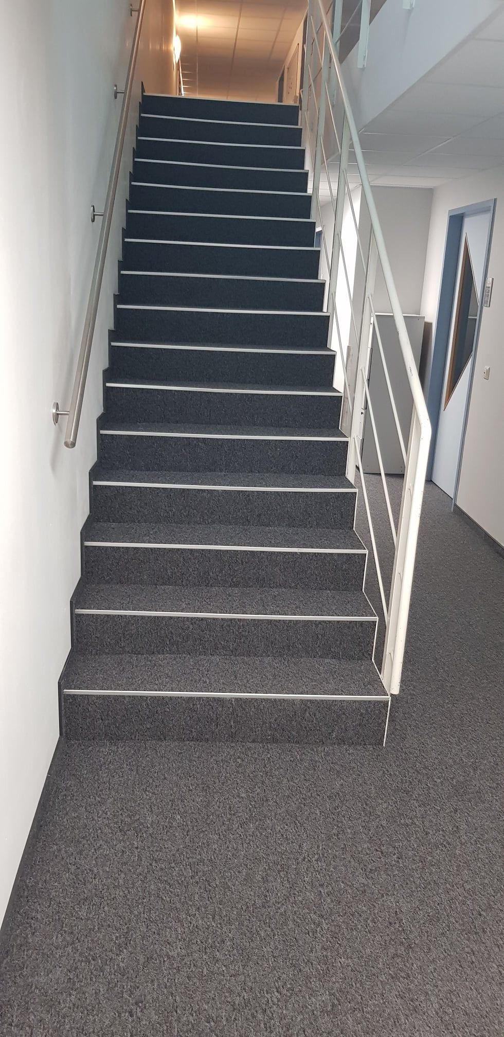 Schody w biurowcu wyłożone szarą wykładziną dywanową z białymi listwami zabezpieczającymi krawędzie stopni, biała balustrada.