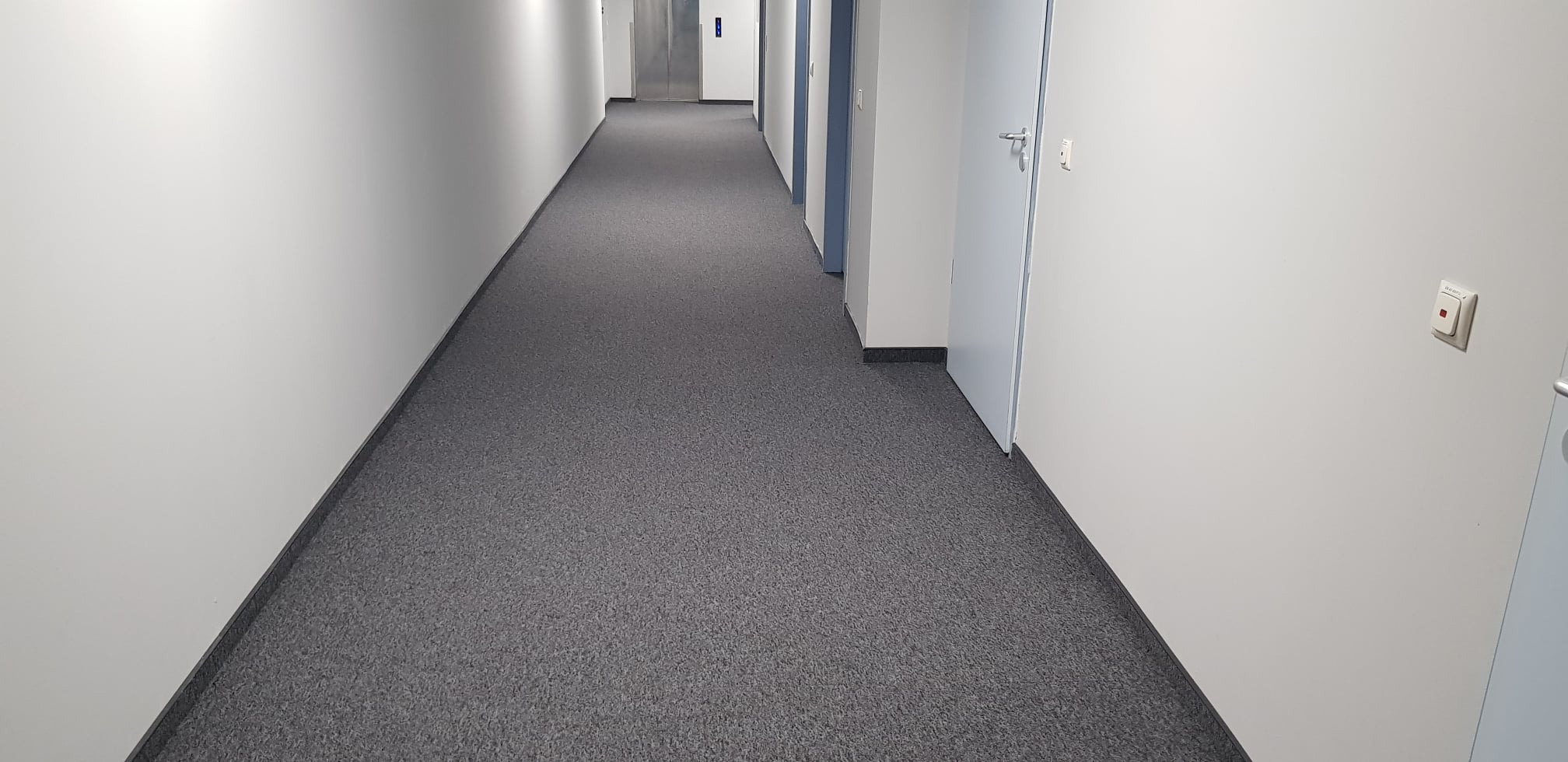 Długi korytarz biurowy z szarą wykładziną dywanową, białymi ścianami i drzwiami, widoczne oświetlenie sufitowe i wyłącznik światła na ścianie.