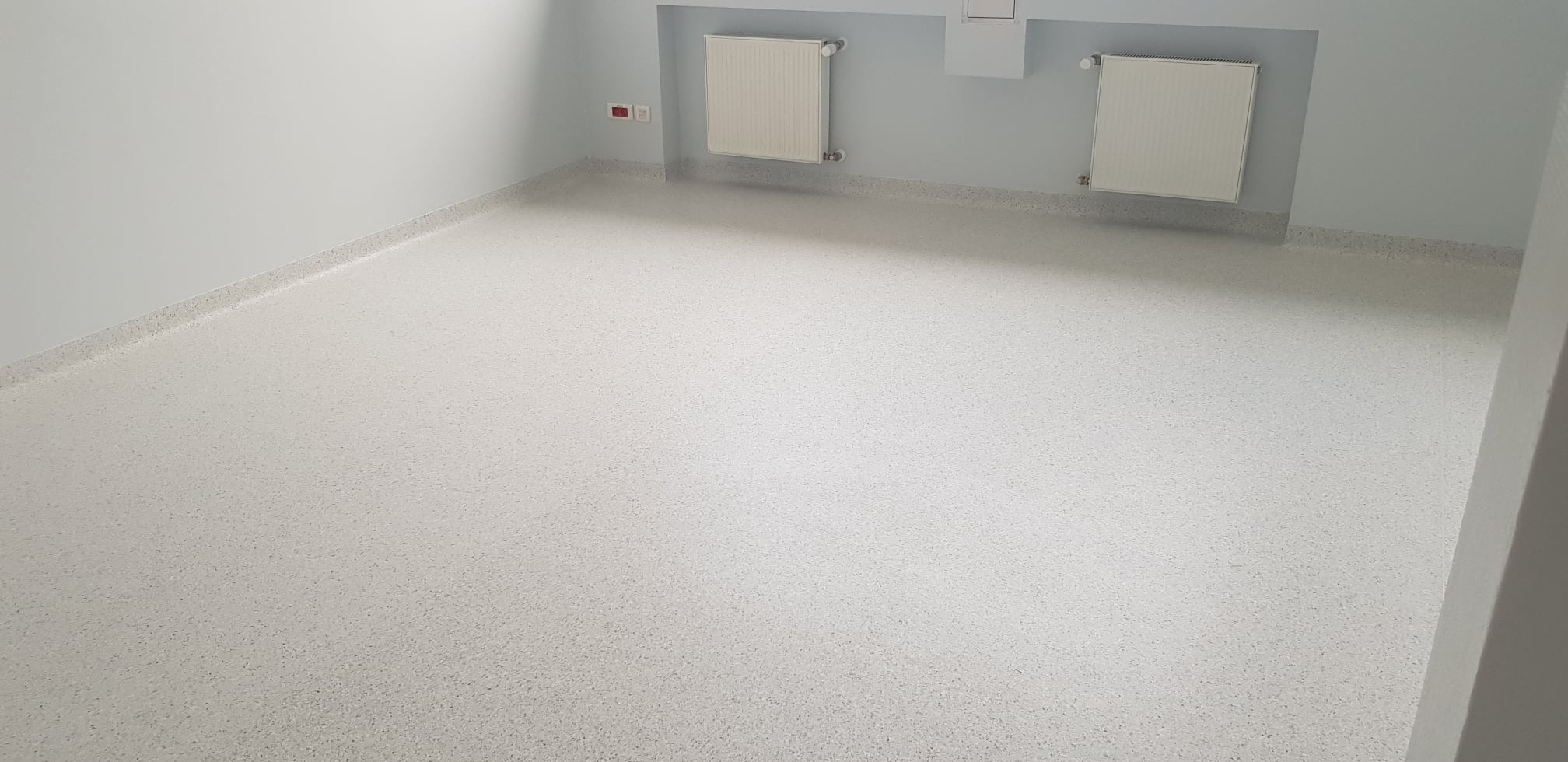 Jasne, puste pomieszczenie z grzejnikami i podłogą wykończoną jasnym, nakrapianym materiałem PCV.