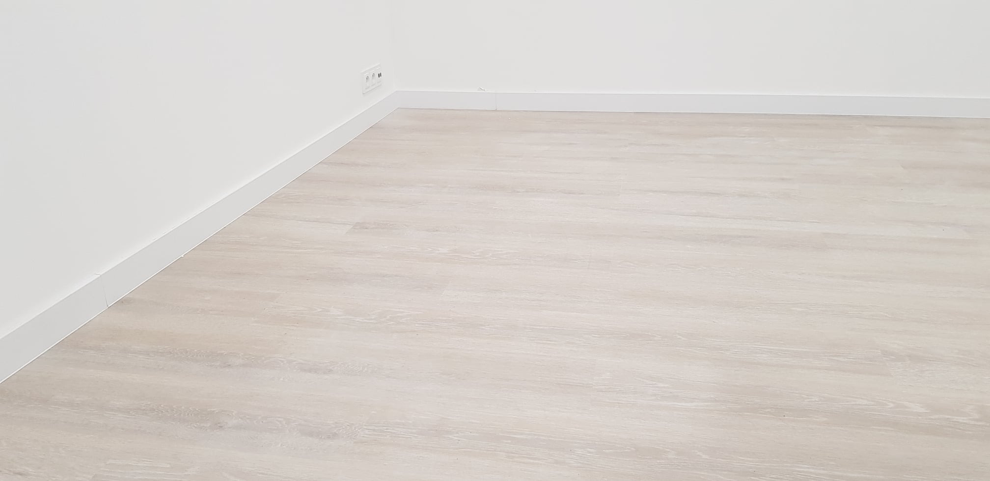 Jasny, drewnopodobny panel podłogowy ułożony w rogu pomieszczenia z białymi ścianami i listwami przypodłogowymi, widoczne gniazdko elektryczne.