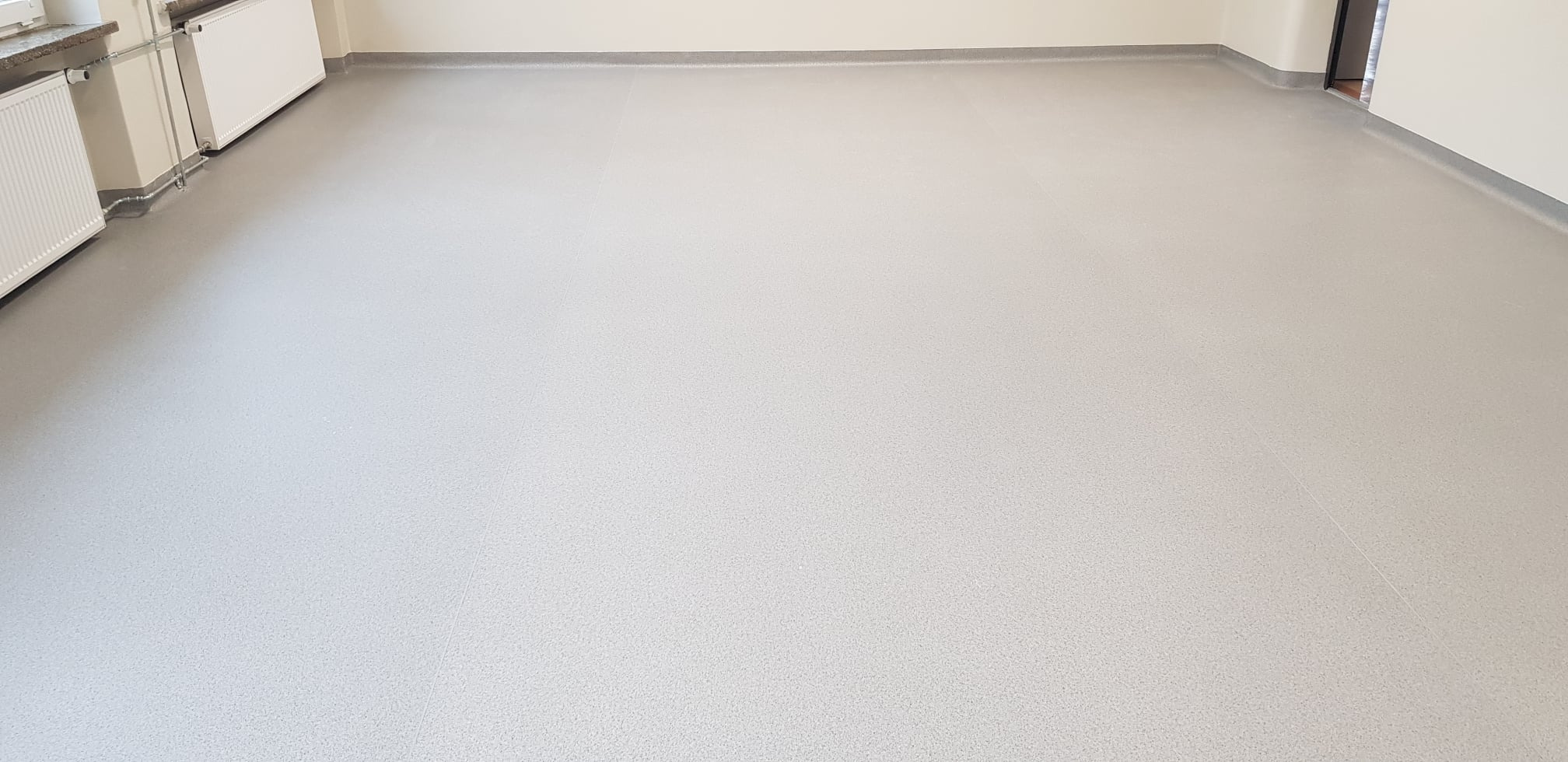 Jasnoszara, gładka podłoga z drobnym wzorem, listwy przypodłogowe w kolorze szarym, grzejniki na ścianie po lewej stronie, jasne ściany, widoczne łączenia paneli podłogowych.