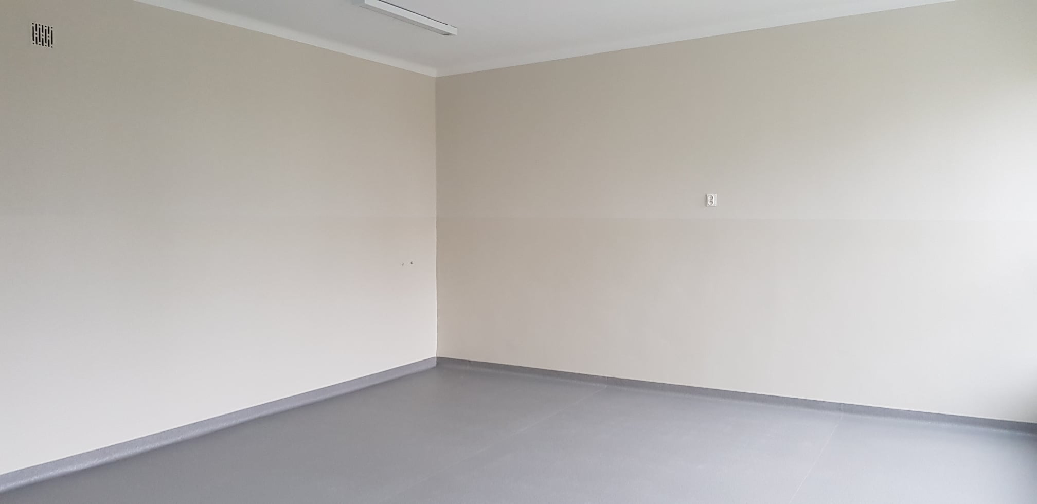 Pomalowane na jasny beż narożne wnętrze z szarą podłogą i białą listwą przypodłogową, widoczny włącznik elektryczny i kratka wentylacyjna.