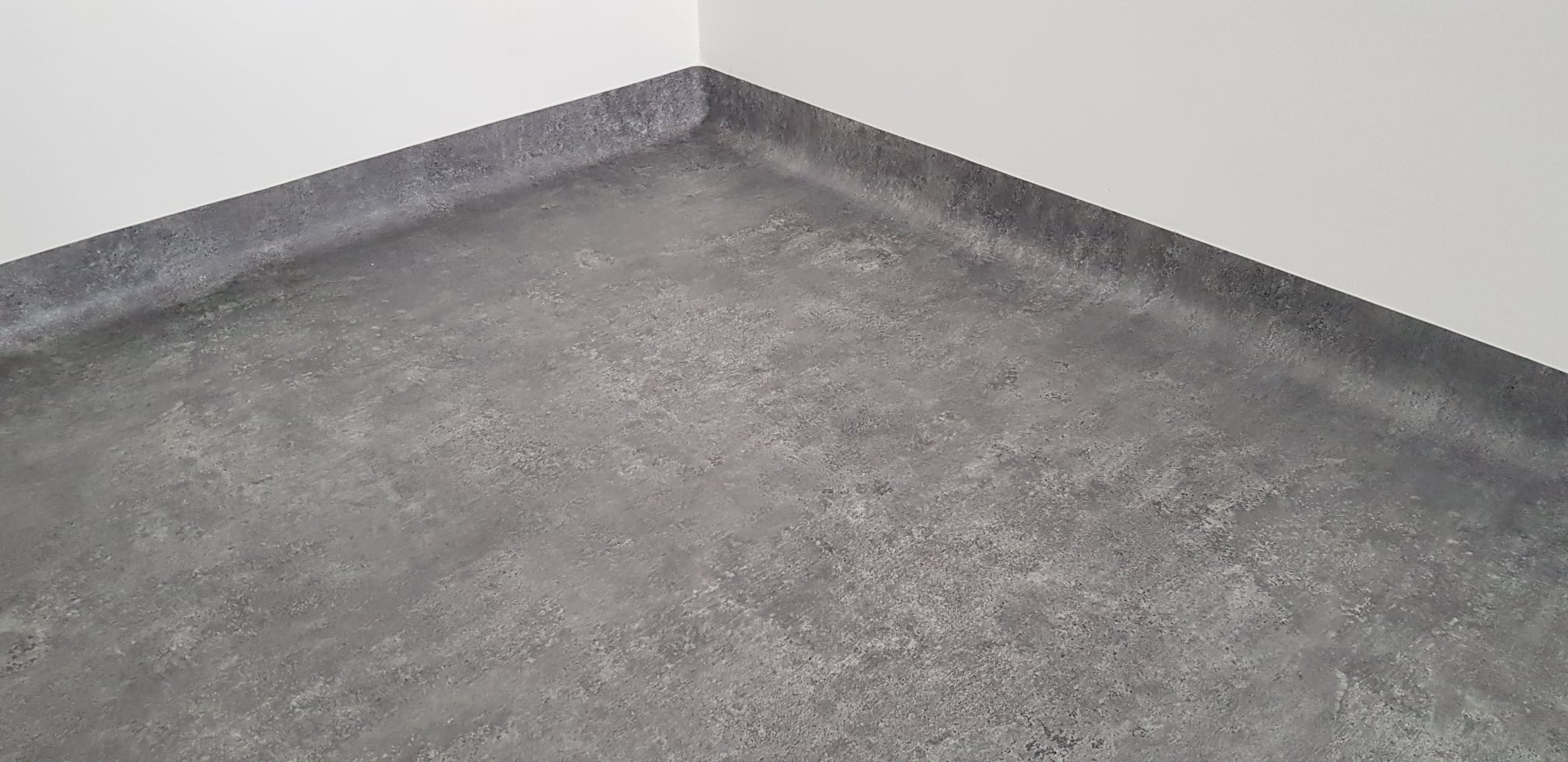 Wykończona szara podłoga w rogu pomieszczenia z białymi ścianami, widoczne połączenie podłogi ze ścianą.