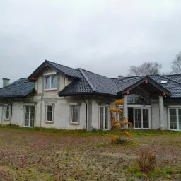 Dom w trakcie budowy z czarną dachówką i widoczną więźbą dachową od frontu, na działce z opadłymi liśćmi, pochmurny dzień.