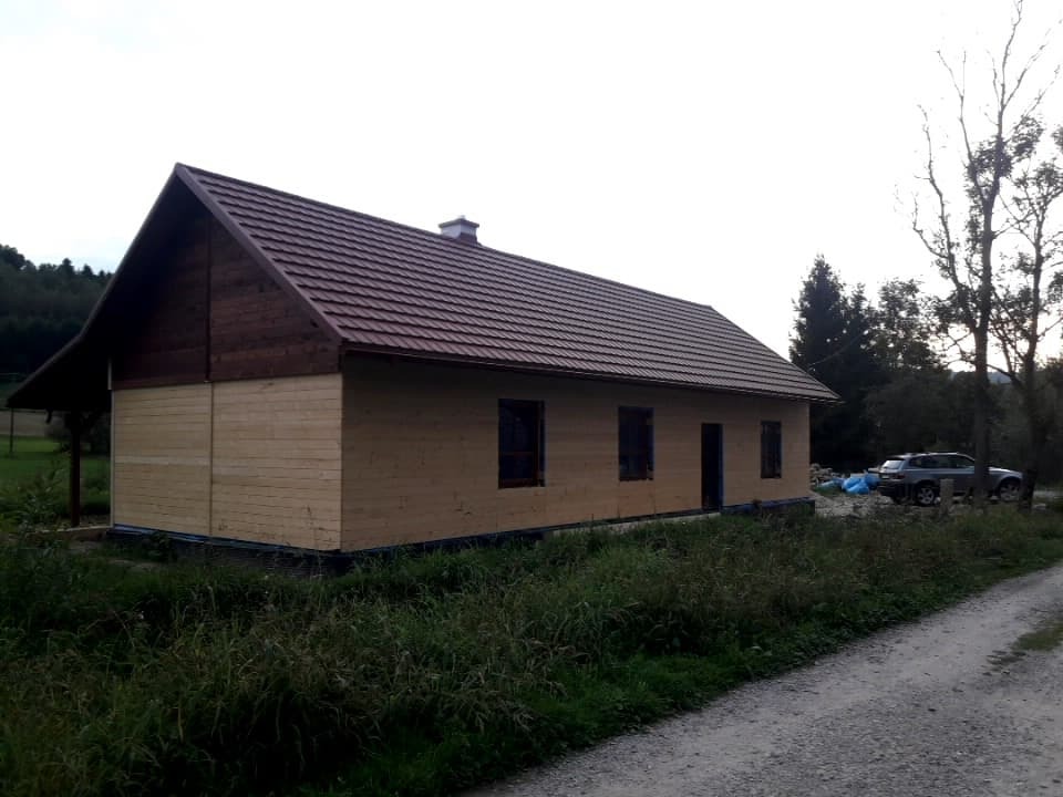 Niski parterowy dom z drewnianą elewacją i nowym dachem z brązowej blachodachówki, widok z boku, w tle las i samochód.