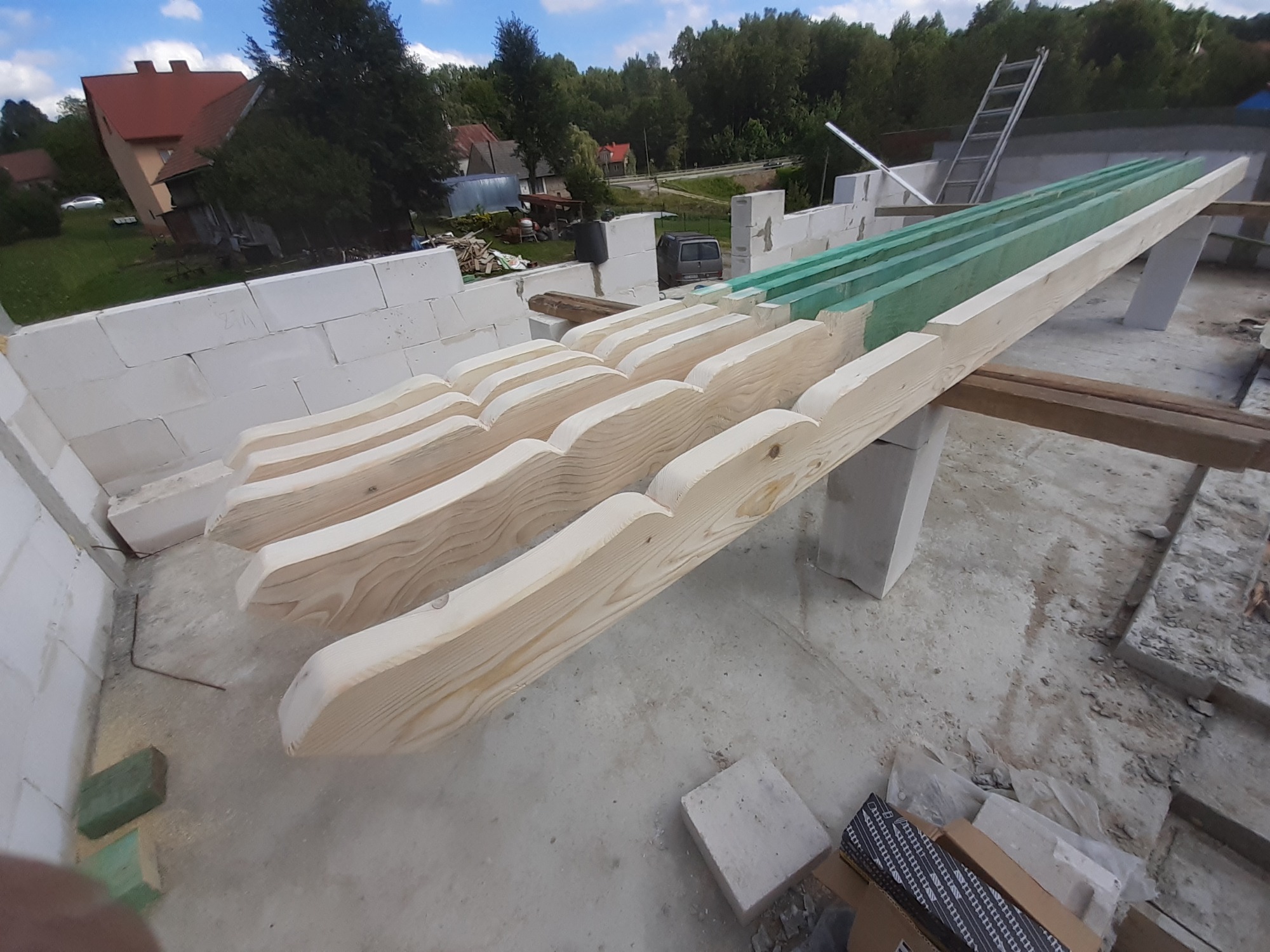 Elementy drewniane ozdobnej konstrukcji dachu, przygotowane do montażu na budowie, widoczne bloczki betonowe i drabina w tle.